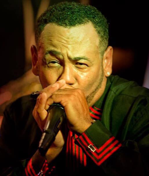 Man wearing black with red trim plays harmonica