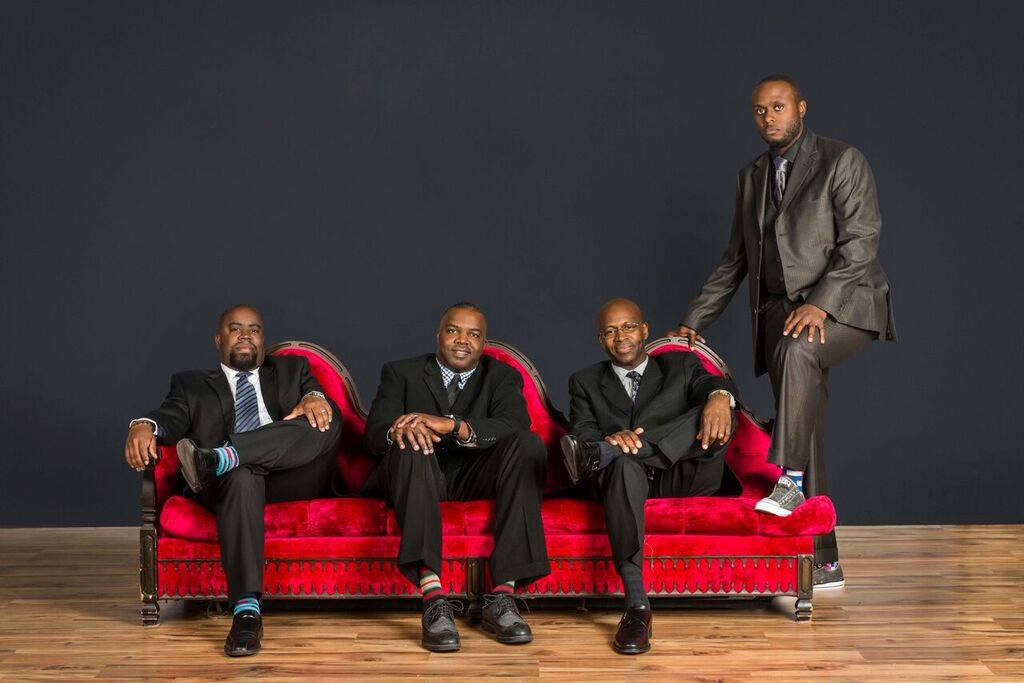 Four men sitting on a red velvet sofa with a black wall behind.