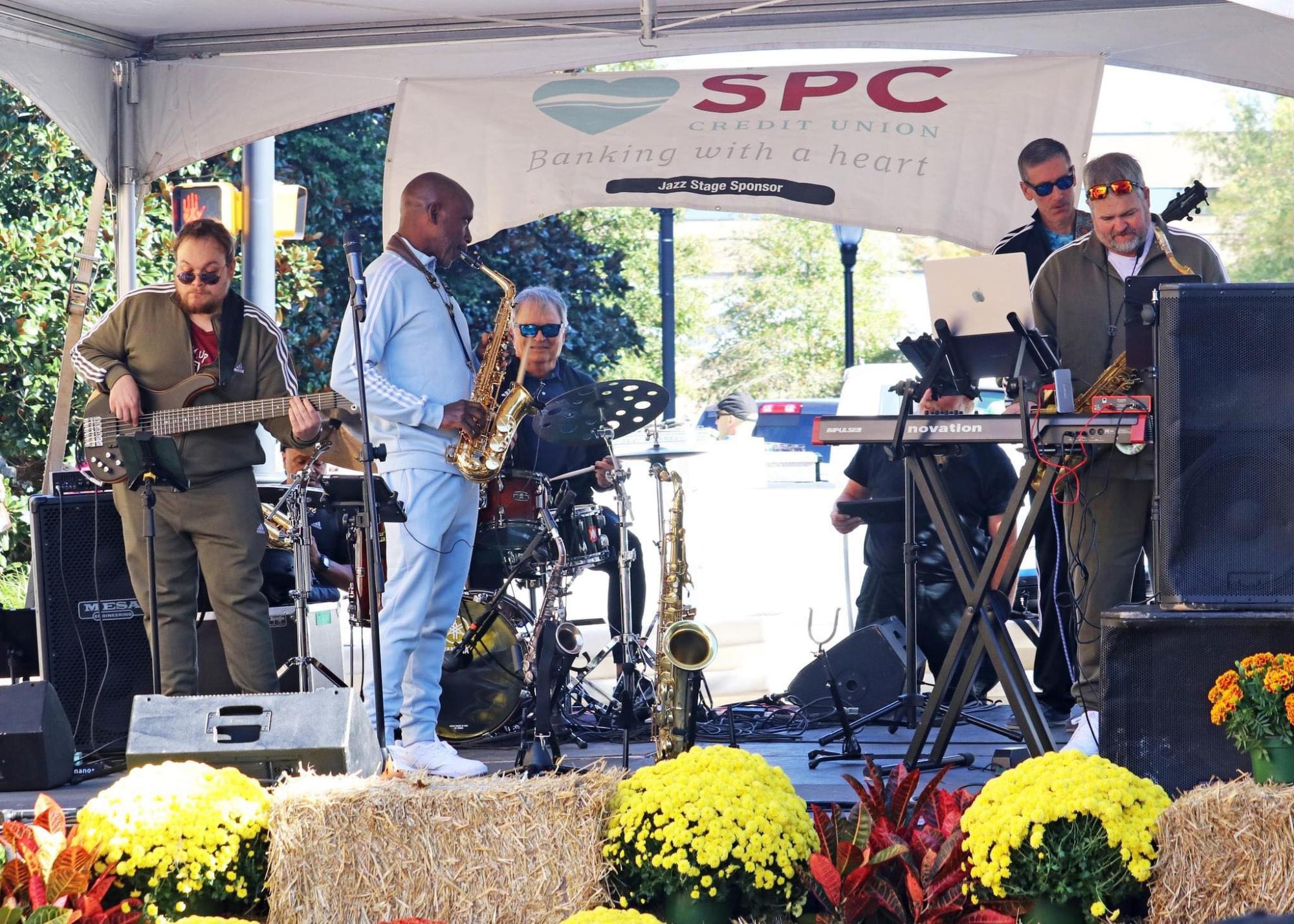 saxophonist in blue suit plays with four other musicians on a stage with flowers in front