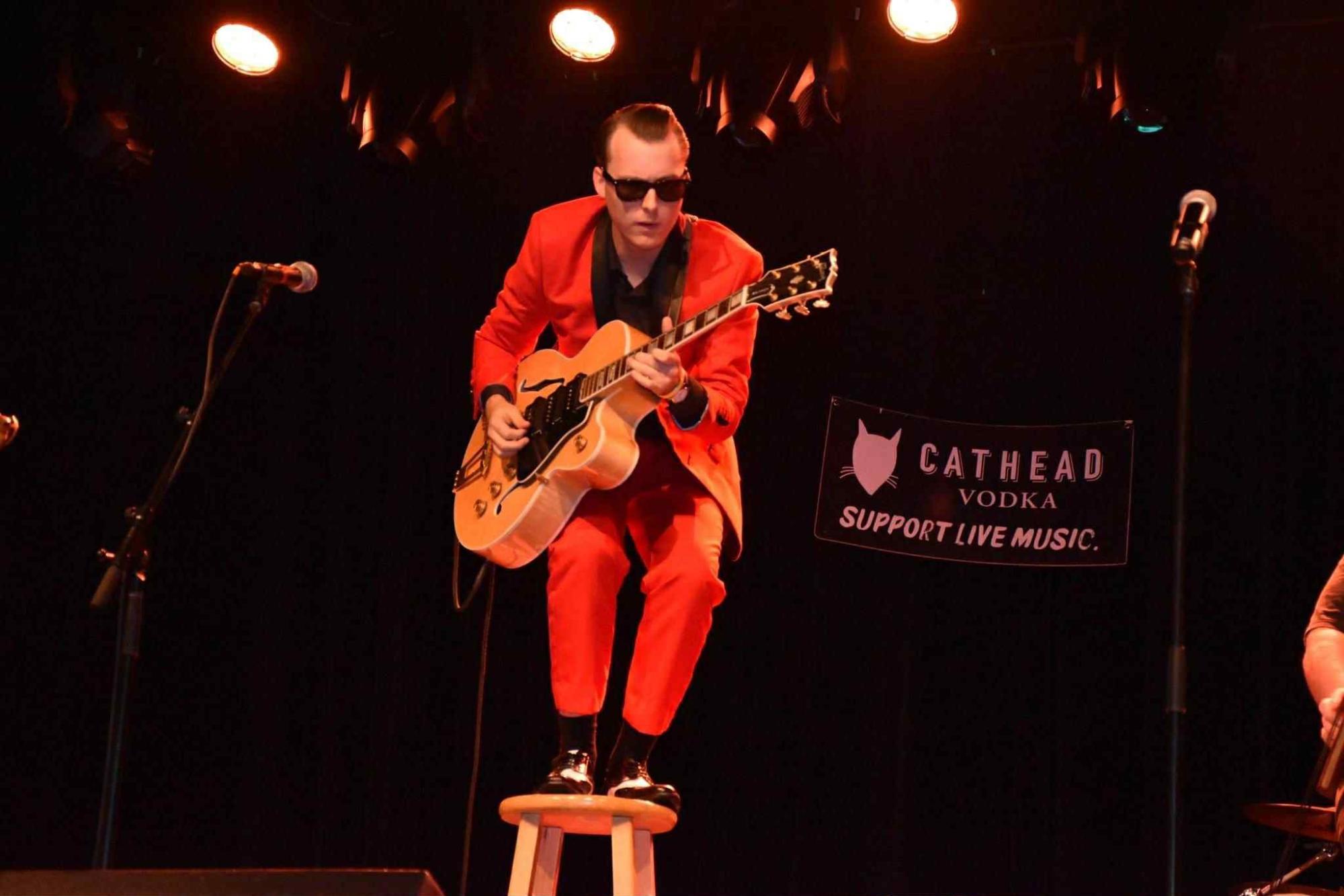 A musician playing blues guitar stands on a stool wearing a red suit.