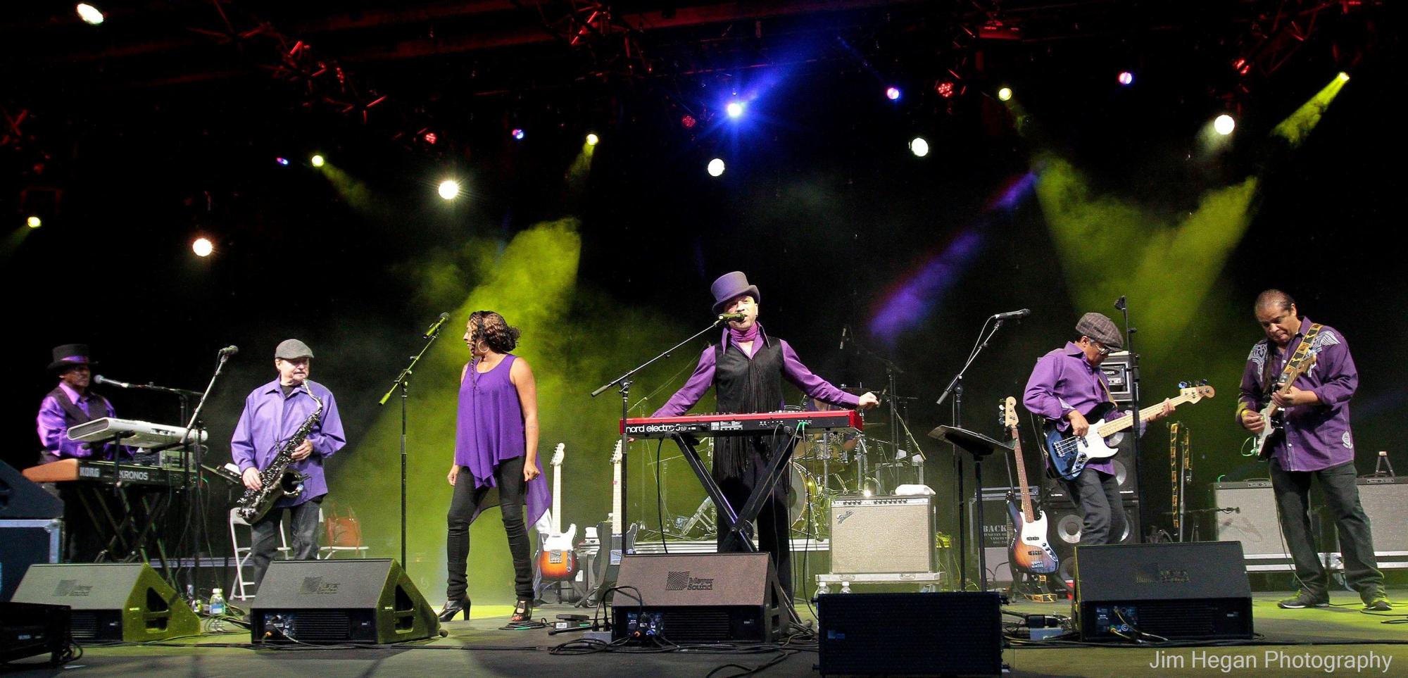 Six band members dressed in purple play on a stage. The center man is in a top hat.